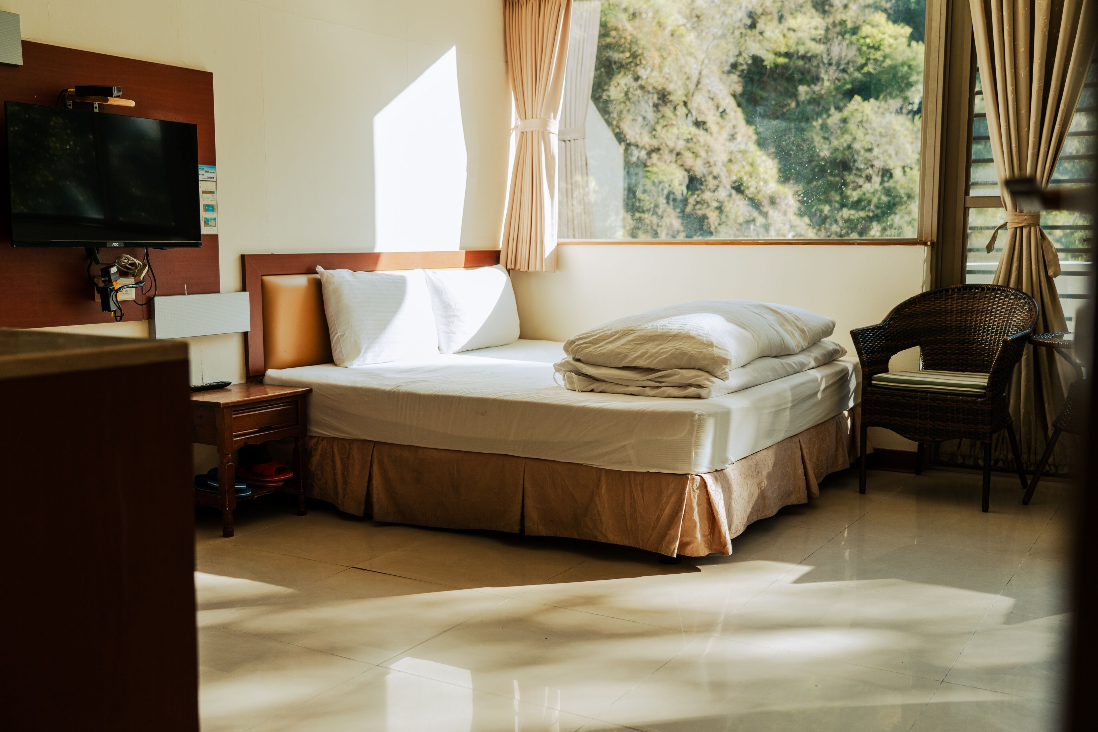 A hotel room with a bed, a television, a window with curtains, and a chair.