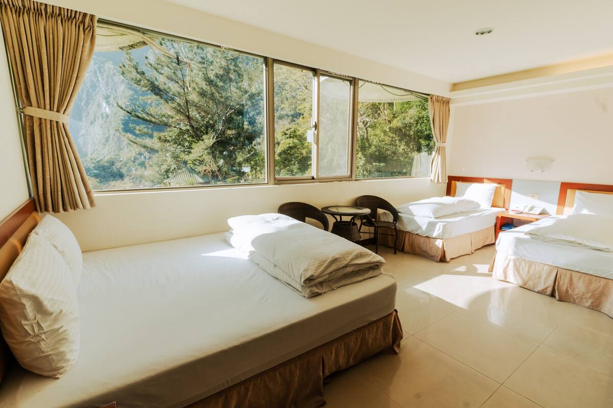 A hotel room with two beds, a small table, and a view of trees through large windows.