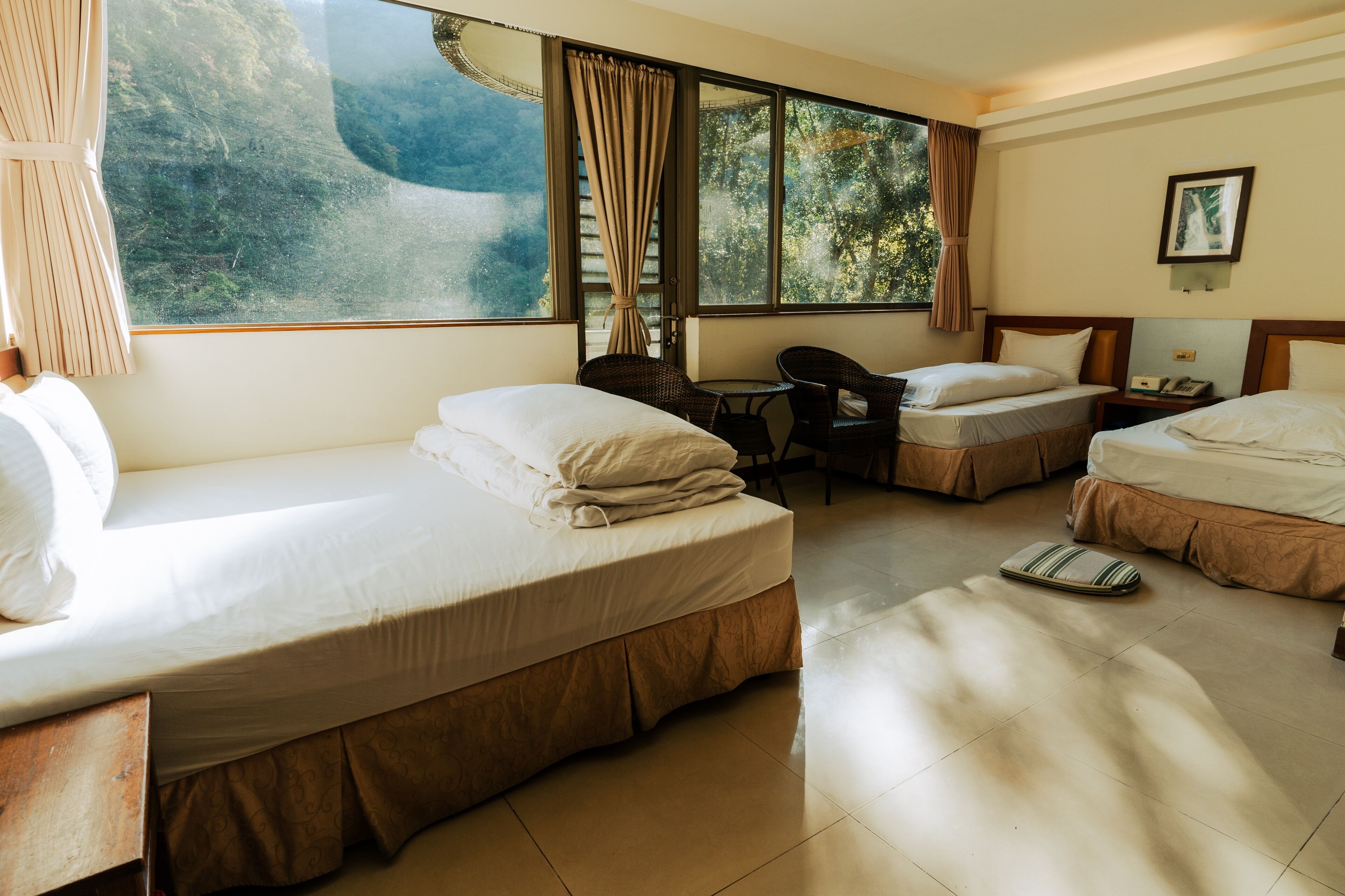 A hotel room with two beds, a small table, a chair, and a window with a view of a mountainous landscape.