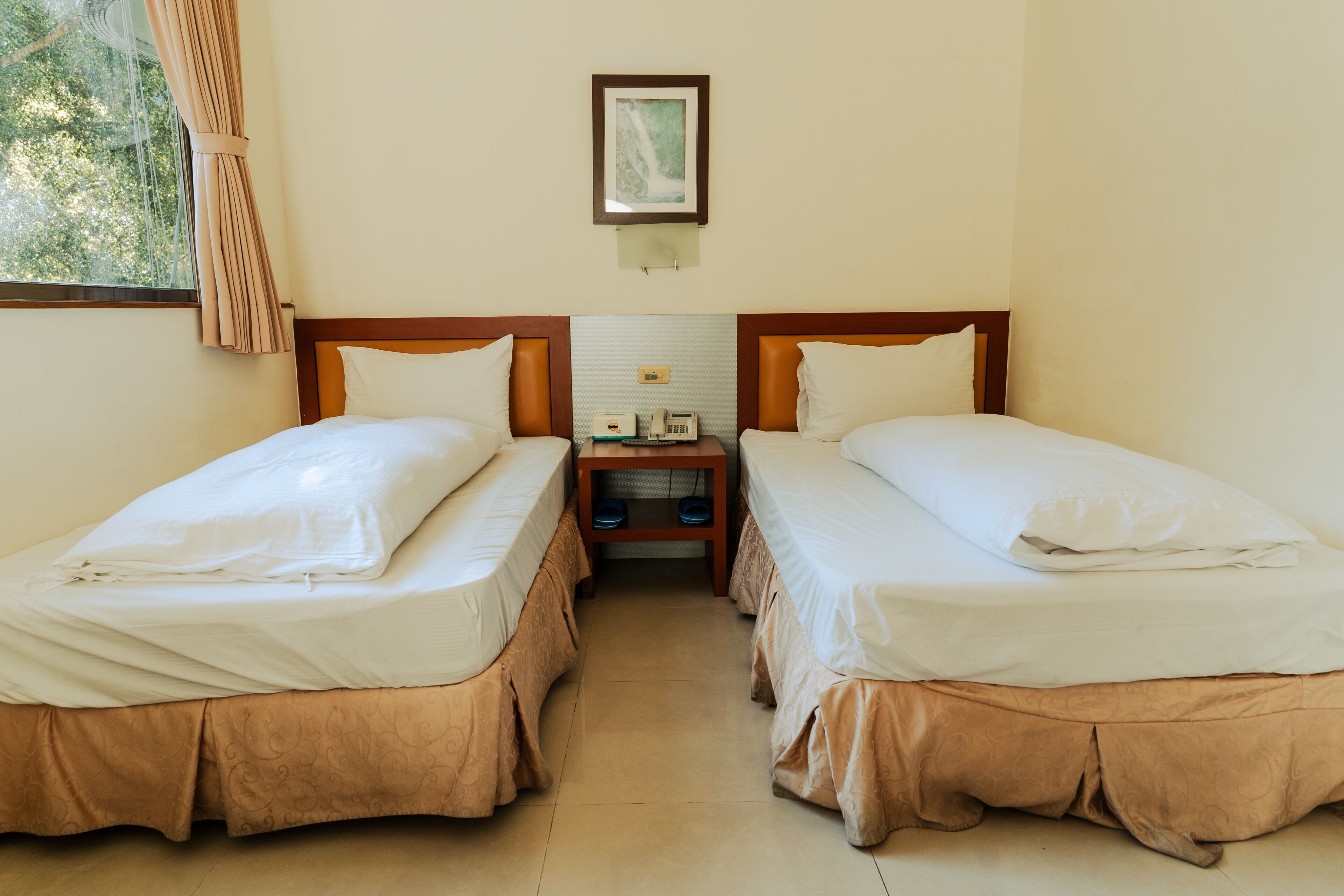 A hotel room with two single beds, a nightstand with a phone and a small table, and a framed picture on the wall.