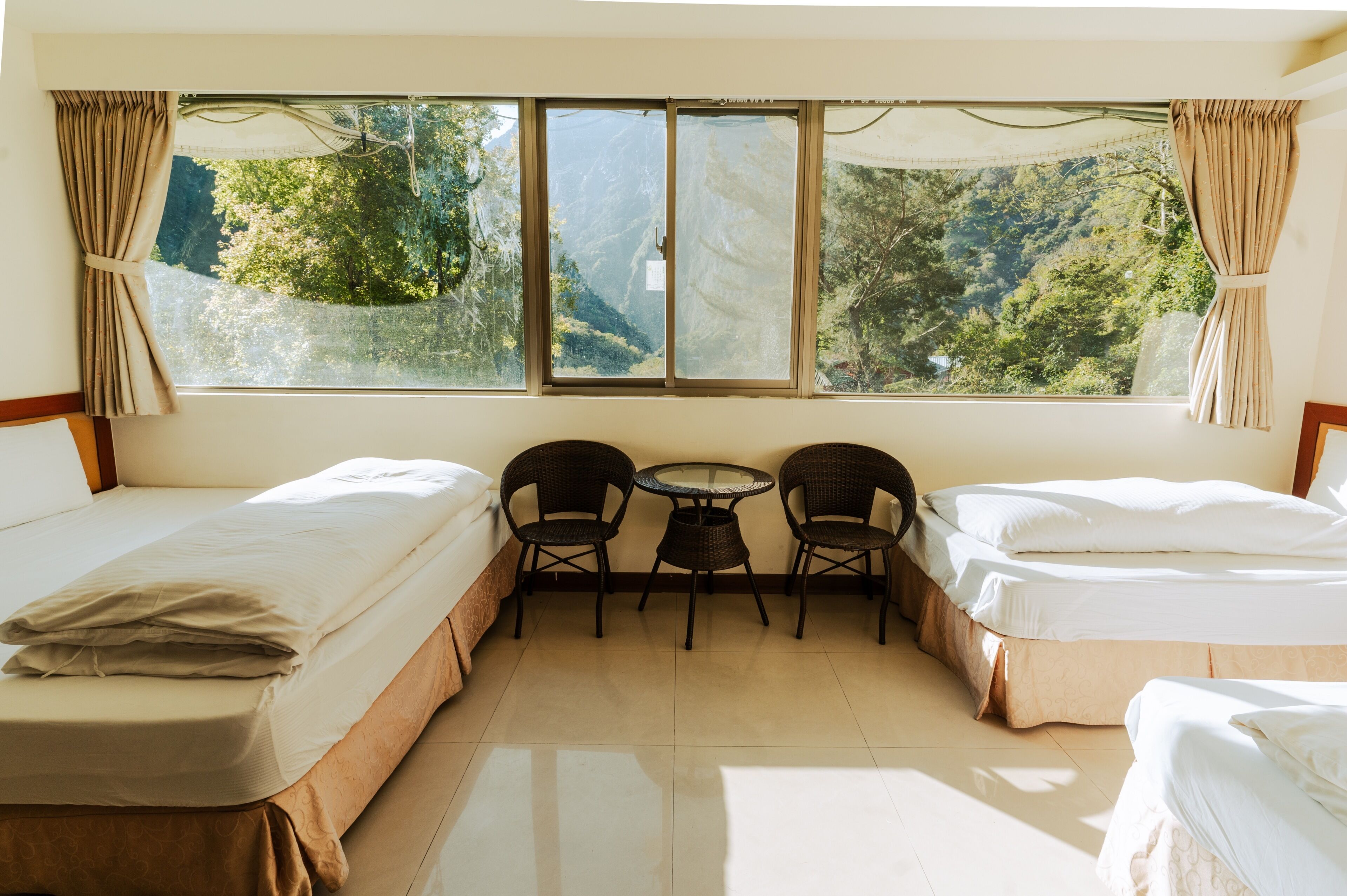 Una habitación con dos camas, una mesita y sillas, y un ventanal que ofrece vistas a árboles y montañas.