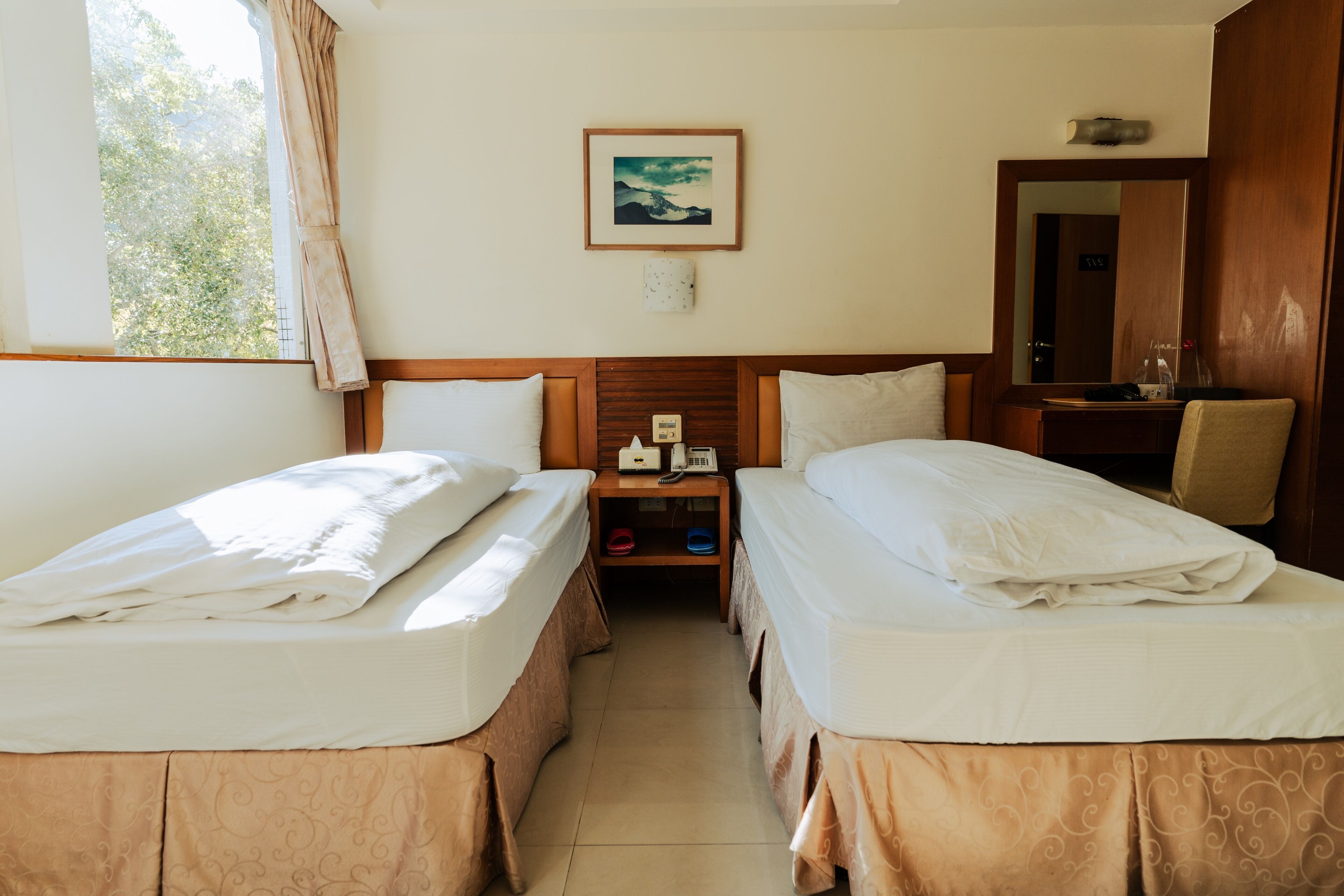 A hotel room with two beds, a nightstand, a mirror, and a window with curtains.