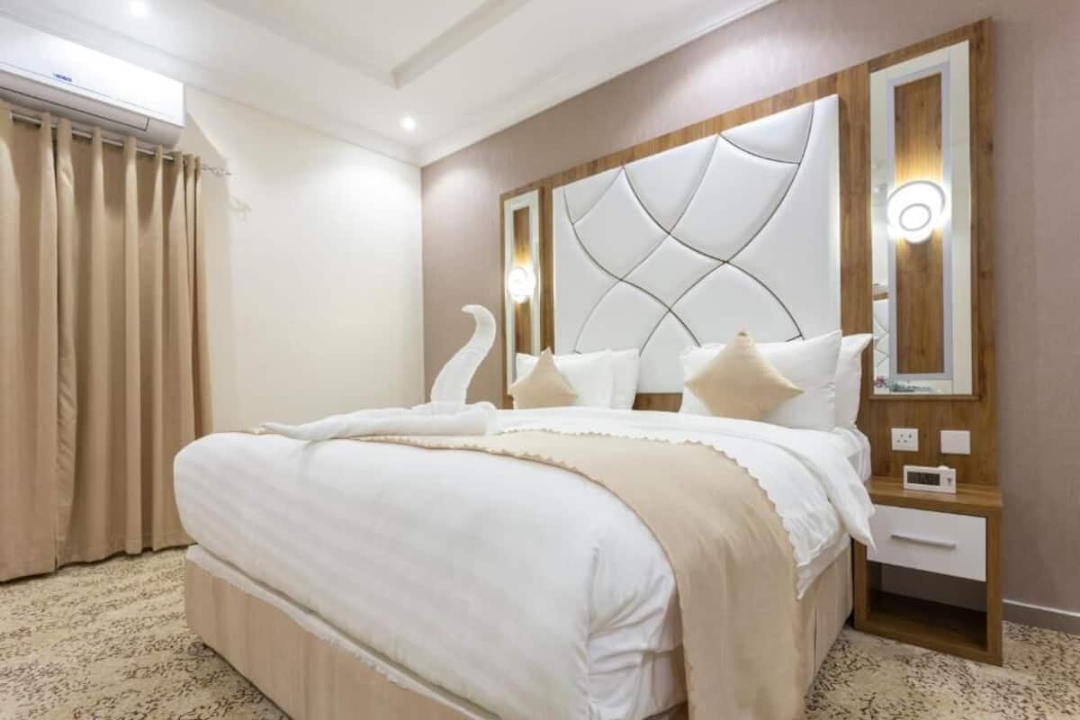 Un dormitorio con una cama grande, cabecera con diseño geométrico, mesita de noche con un reloj y un espejo con apliques de pared.