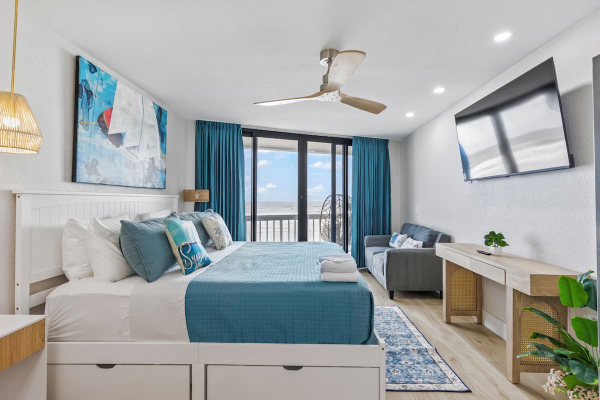 A modern hotel room with a bed, a desk, a TV, and a balcony with a view.
