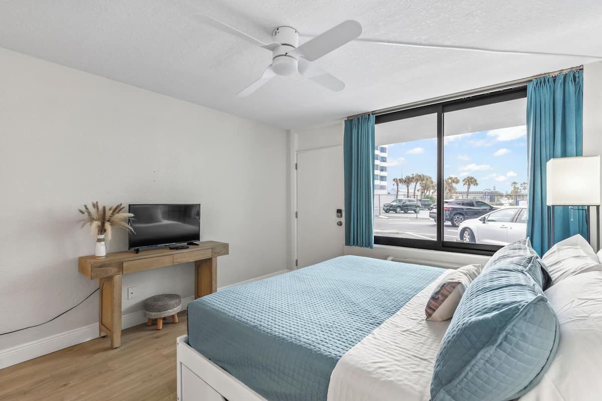 A bedroom with a bed, a TV on a wooden stand, a ceiling fan, a window with curtains, and a view of the street.