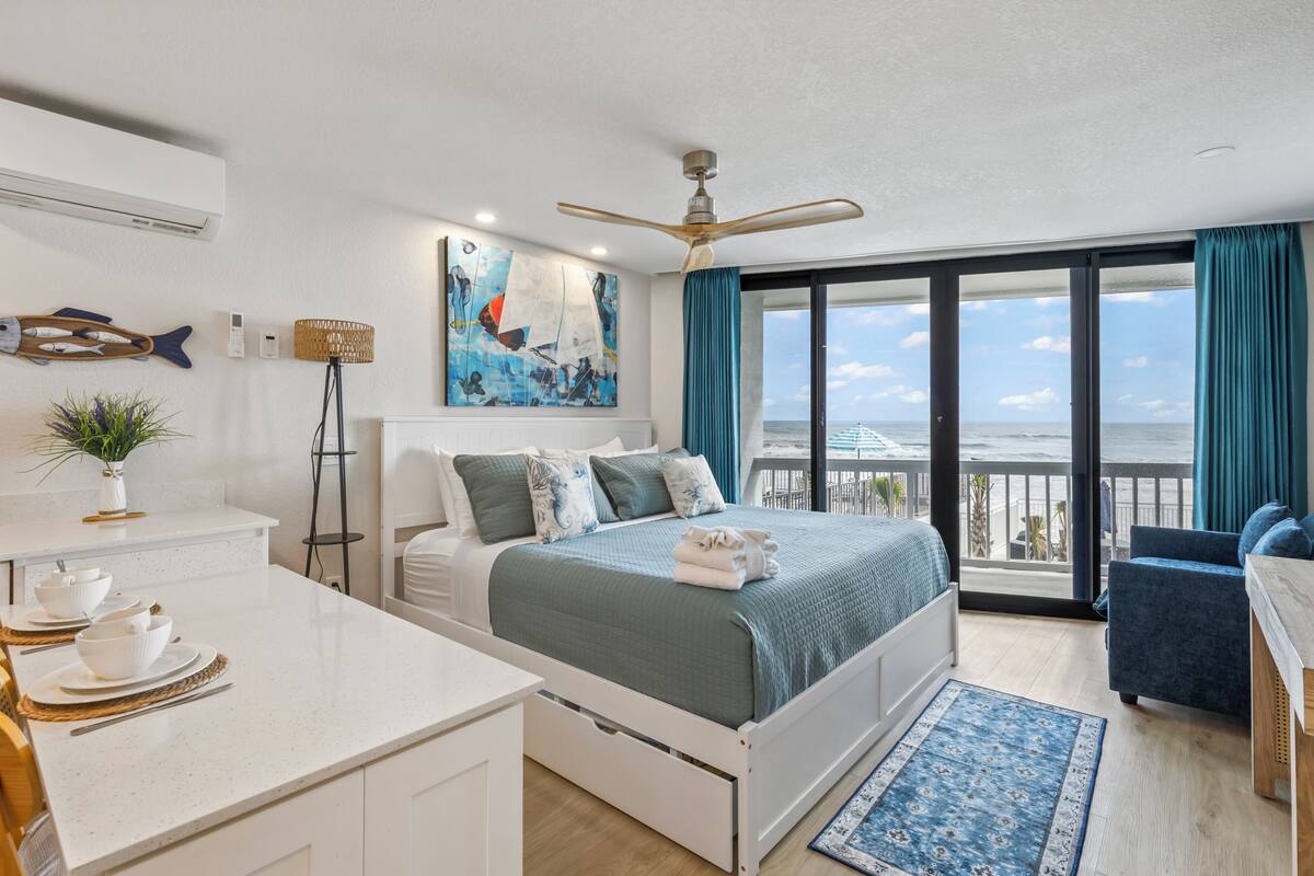 A modern bedroom with a bed, a desk, a chair, a balcony with a view, and a ceiling fan.