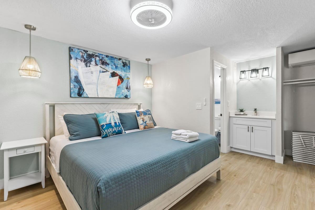 A bedroom with a bed, bedside tables, a ceiling fan, a painting, and a bathroom with a mirror and cabinets.