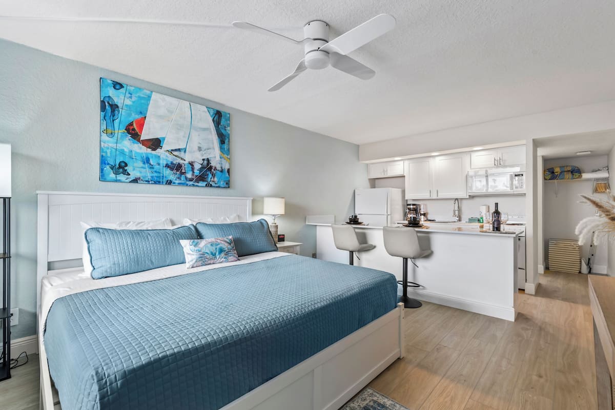 A modern bedroom with a large bed, a ceiling fan, and a kitchen area visible through an open door.
