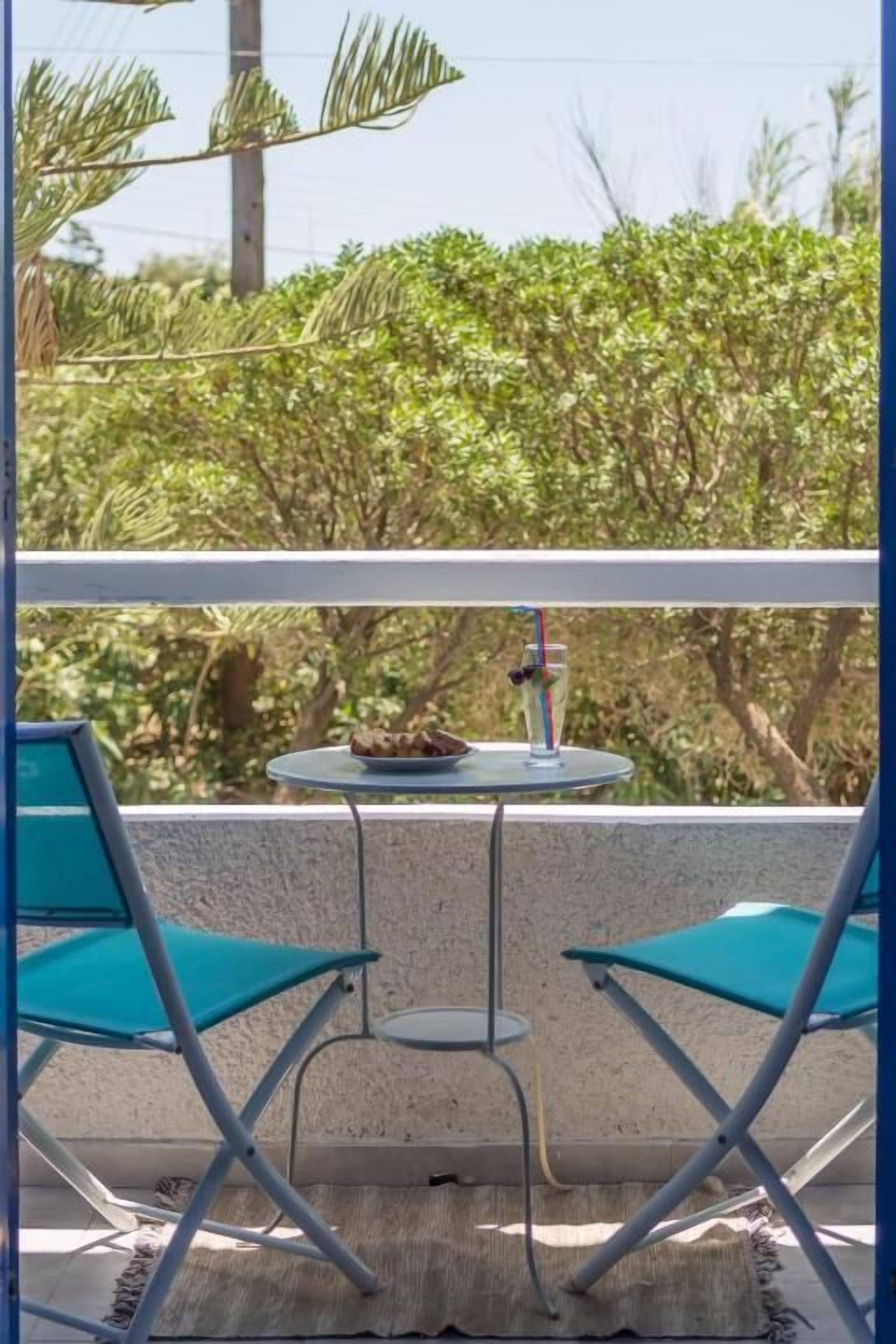 Un balcón con dos sillas turquesas, una mesita redonda con comida y una bebida, y vistas a vegetación exuberante.