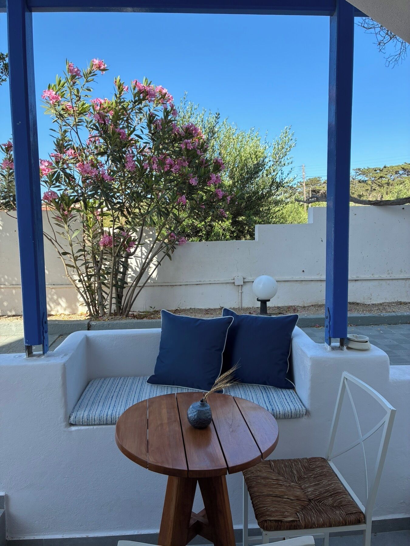 A covered outdoor seating area with a white sofa, two blue cushions, a round wooden table, and a wicker chair.