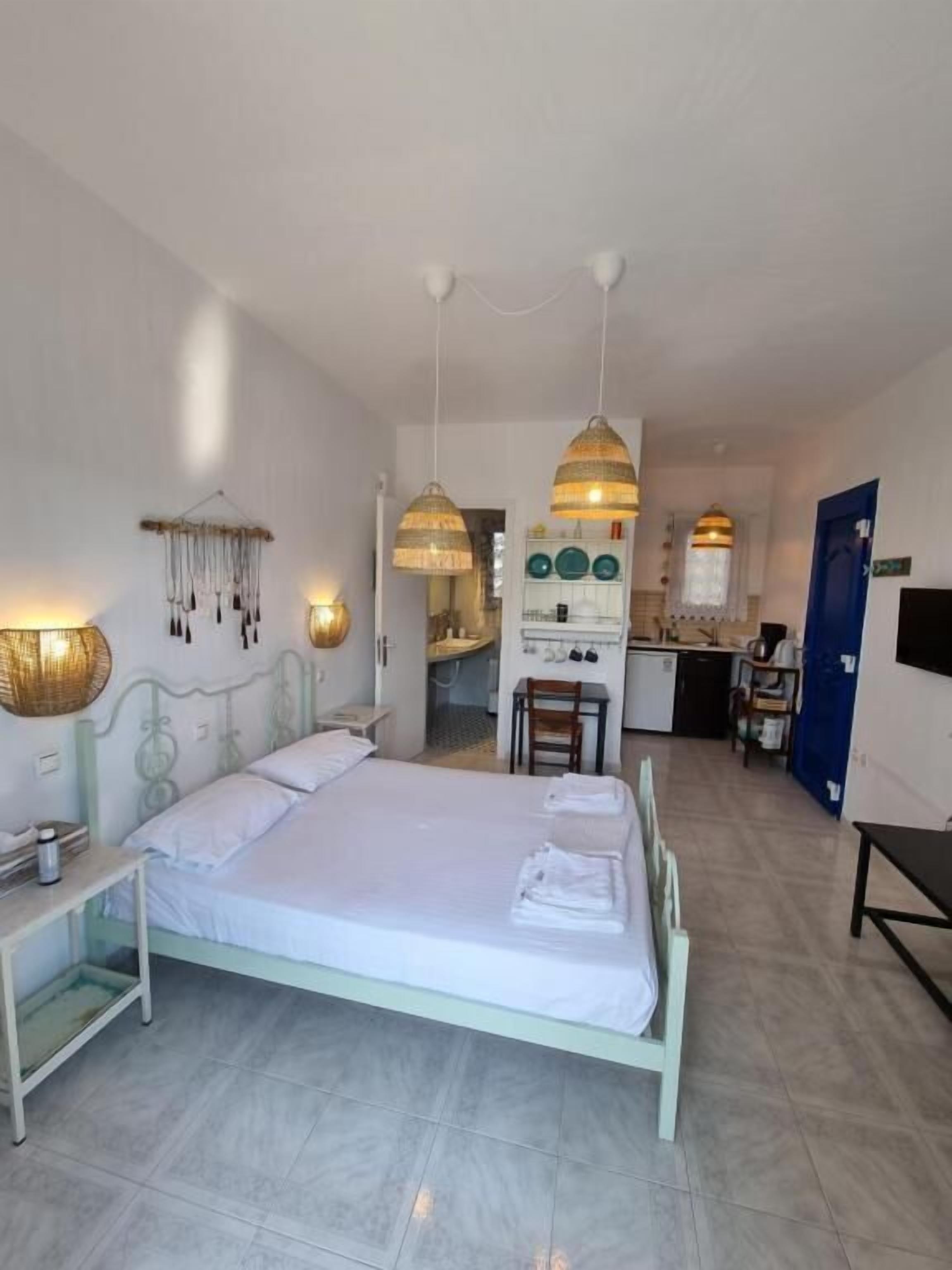 Una habitación de hotel con cama, mesita de noche, lámparas colgantes y una kitchenette al fondo.