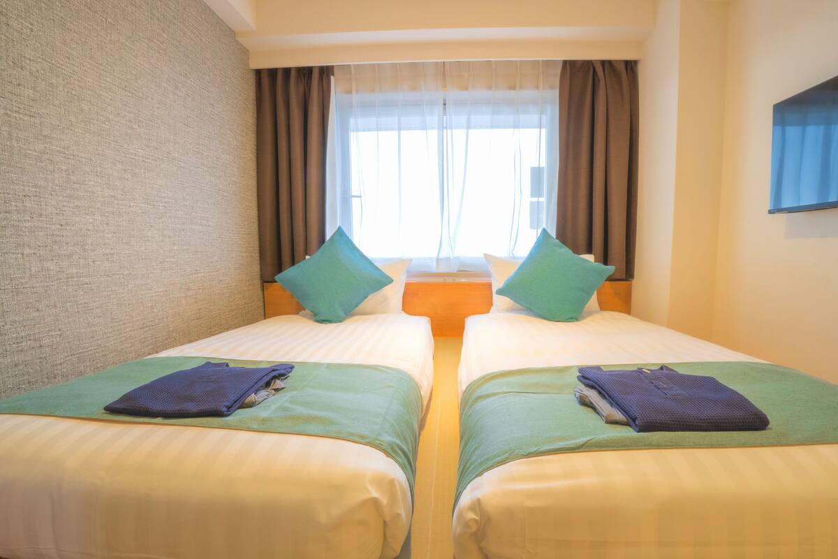 A twin room with two beds, each with a green bedspread and pillows, a window with curtains, and a flat-screen TV mounted on the wall.