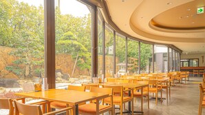 Un vaste espace dédié à la salle à manger, avec de longues tables et des chaises, de grandes fenêtres donnant sur un jardin agrémenté de rochers et d’arbres.