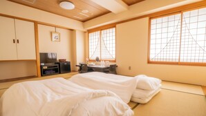 Une pièce de style japonais traditionnel, avec un sol recouvert de tatamis, un lit avec une literie blanche, un bureau avec une chaise, une télévision et une fenêtre à motifs géométriques.
