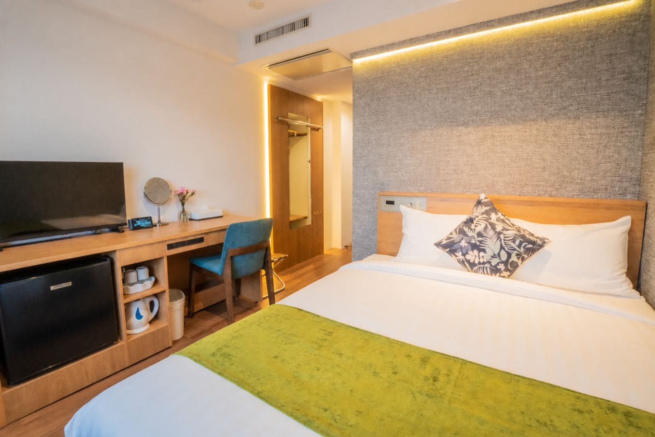 A hotel room with a bed, a desk with a television, a chair, and a small refrigerator.