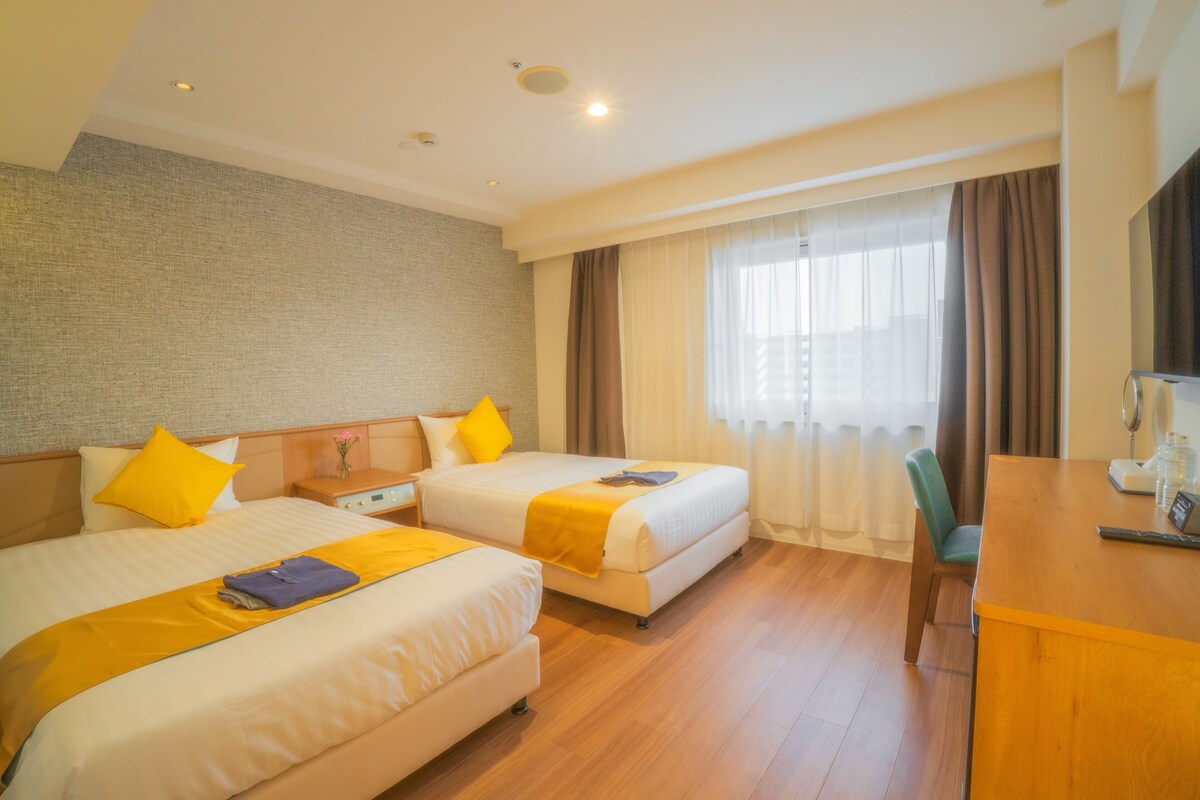Ein Hotelzimmer mit zwei Betten, einem Holztisch und einem großen Fenster mit Vorhängen.