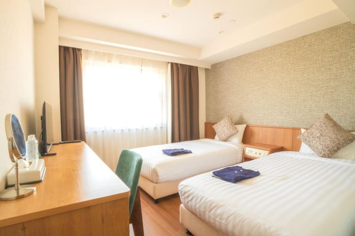 Una habitación de hotel con dos camas, un escritorio con espejo y un ventanal con cortinas.