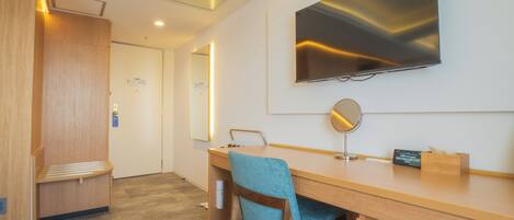 A hotel room with a wooden desk, a chair, a flat-screen TV mounted on the wall, a mirror, and a small wooden box.
