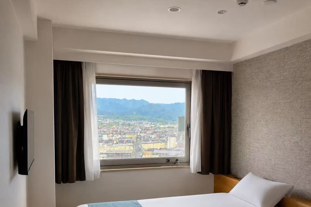 Habitación de hotel con cama, televisión, ventana con cortinas y vistas a la ciudad.
