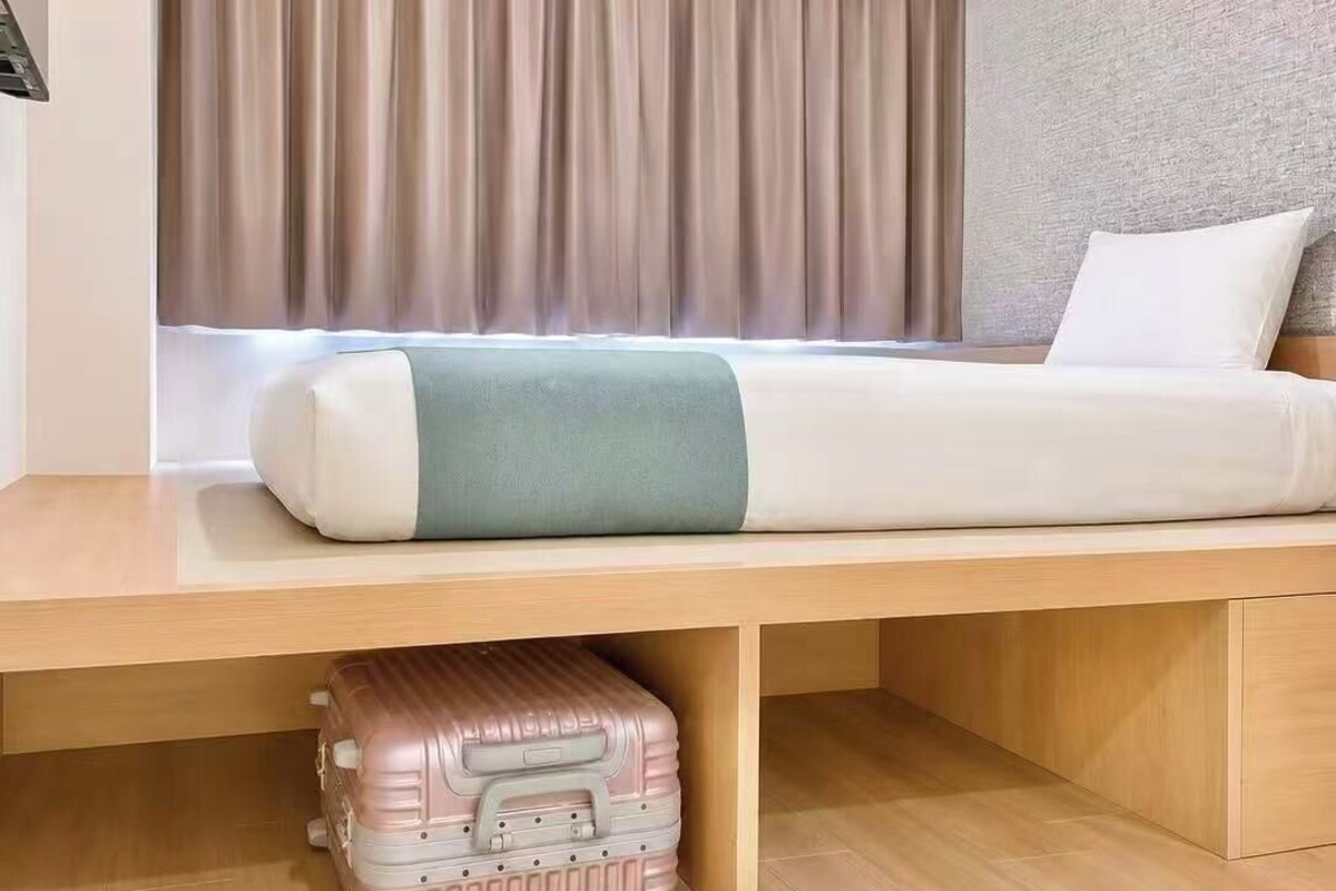 Ein Hotelzimmer mit Bett, Fenster mit Vorhängen, einem Koffer unter dem Bett und einer strukturierten Wand.