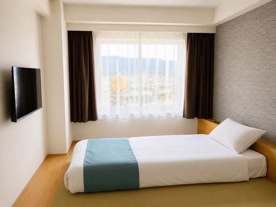 A hotel room with a bed, a television on the wall, a window with curtains, and a view of buildings.