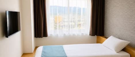 A hotel room with a bed, a television on the wall, a window with curtains, and a view of buildings.