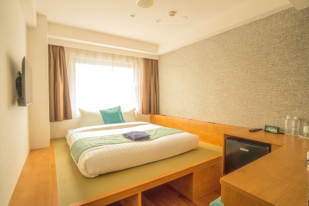 Una habitación de hotel con cama, escritorio, refrigerador y ventana con cortinas.