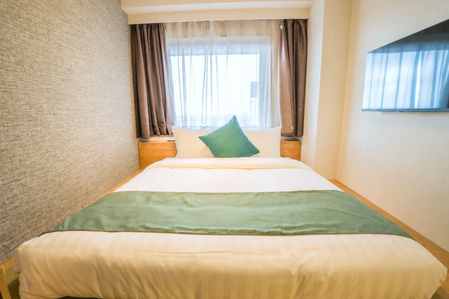 A hotel room with a bed, a nightstand, a window with curtains, and a wall with a textured pattern.