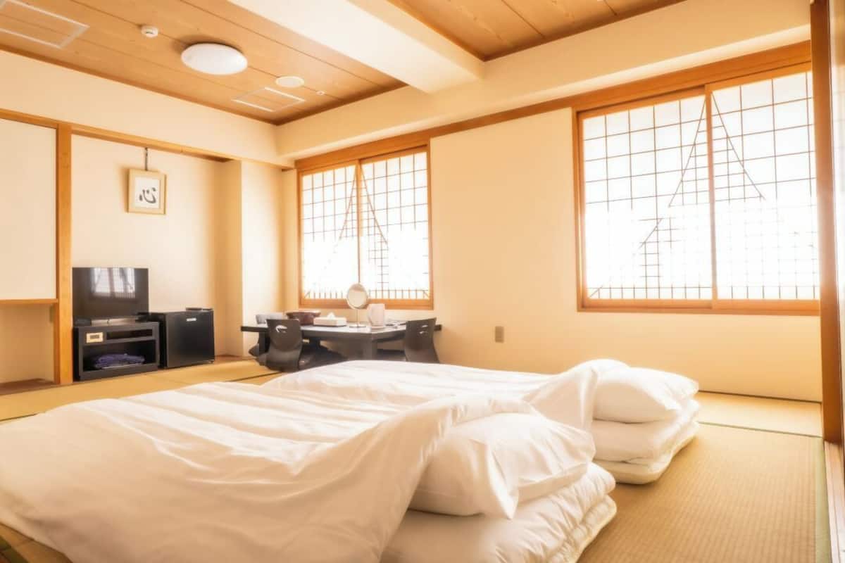 Una habitación tradicional japonesa con suelo de tatami, una cama con ropa de cama blanca, un escritorio con espejo y un televisor.