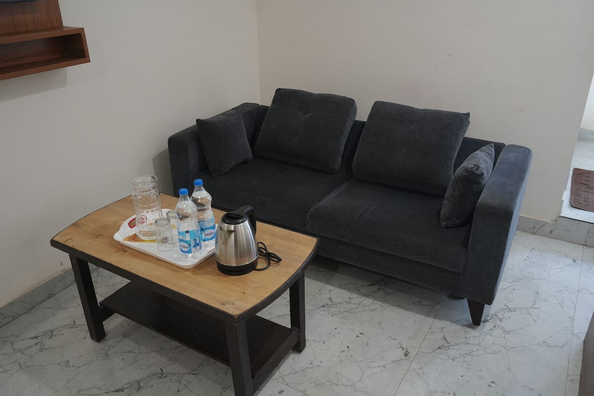 Ein Wohnzimmer mit einem dunkelgrauen Sofa, einem Holztisch mit einer Auflage und verschiedenen Gegenständen, darunter Flaschen und einem Wasserkocher, sowie einem Boden mit Marmorfliesen.