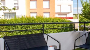Ein Balkon mit blauem Tisch und zwei schwarzen Stühlen, einer weißen Lampe und Blick auf moderne Gebäude im Hintergrund.