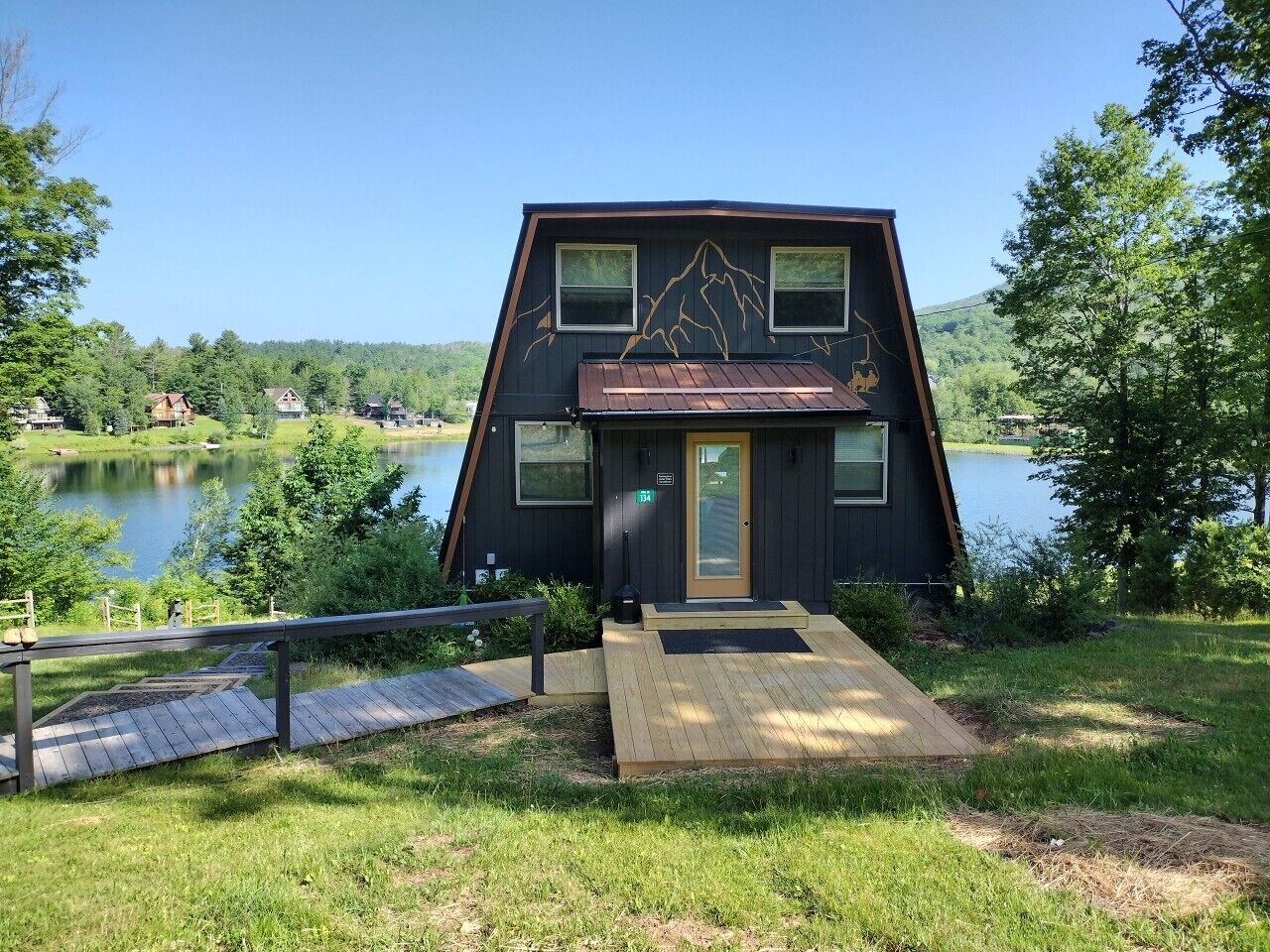 A cabin with a wooden deck, a door, and windows overlooking a lake.