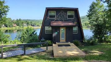 A cabin with a wooden deck, a door, and windows overlooking a lake.