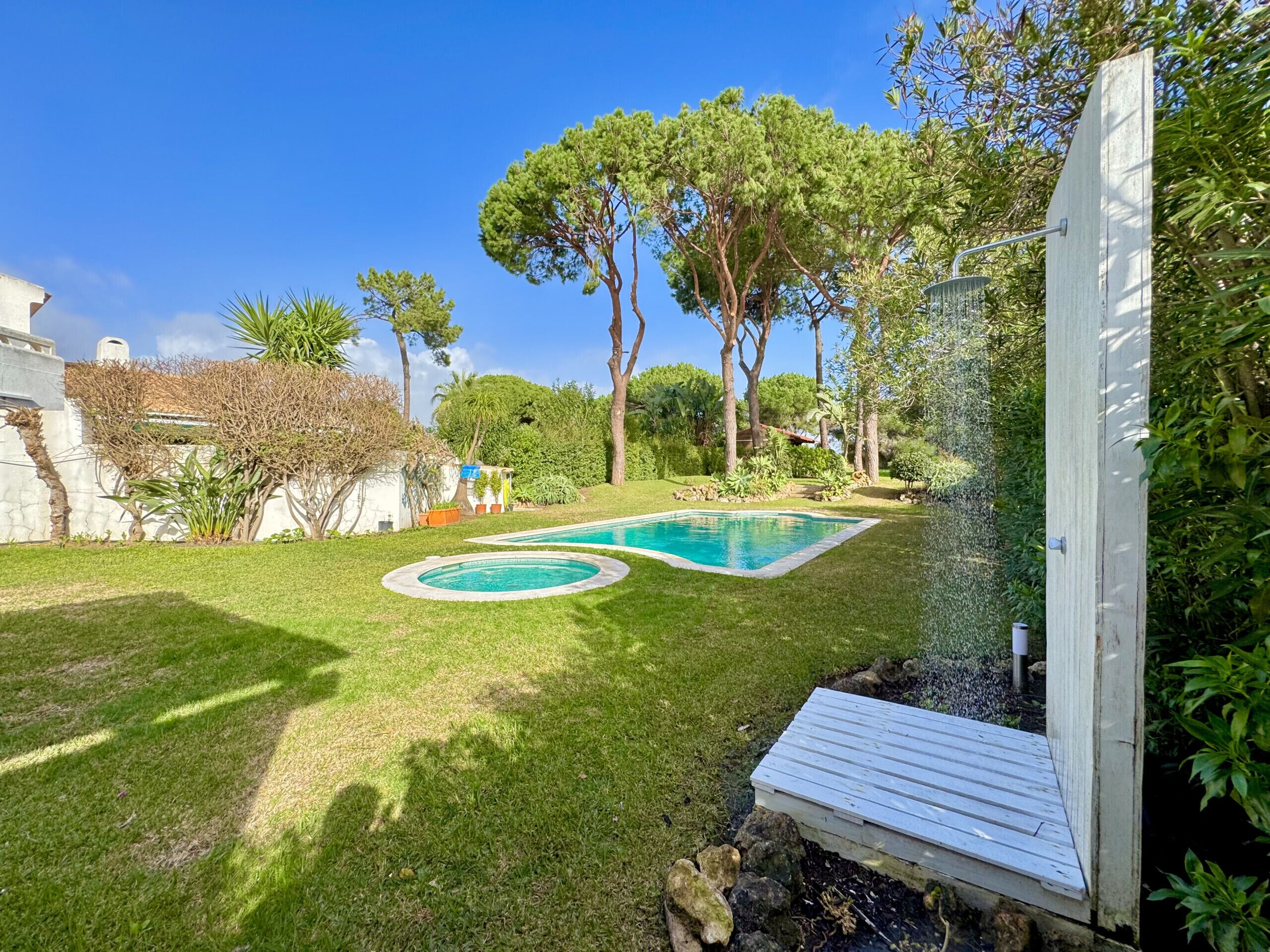 Un patio trasero con piscina, terraza de madera y diversas plantas y árboles.