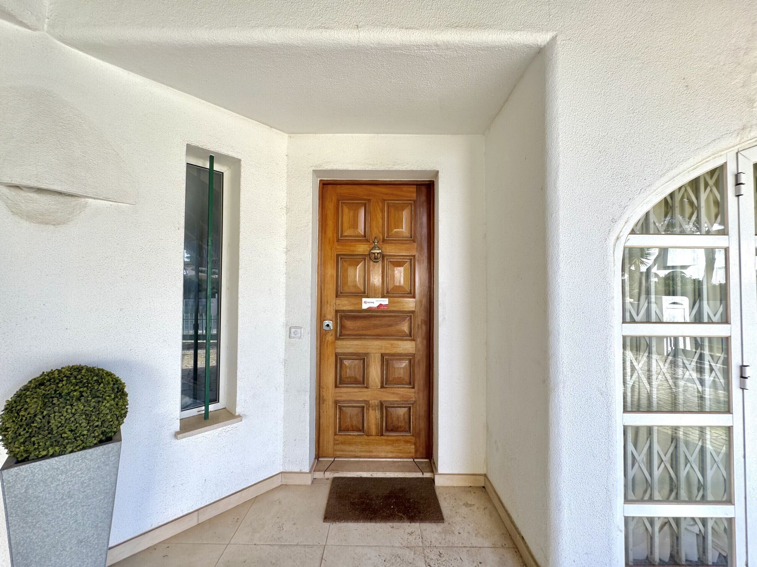 Une porte en bois ornée d’un heurtoir en laiton, une petite fenêtre à cadre vert et un pot de fleurs placé à côté de la porte.