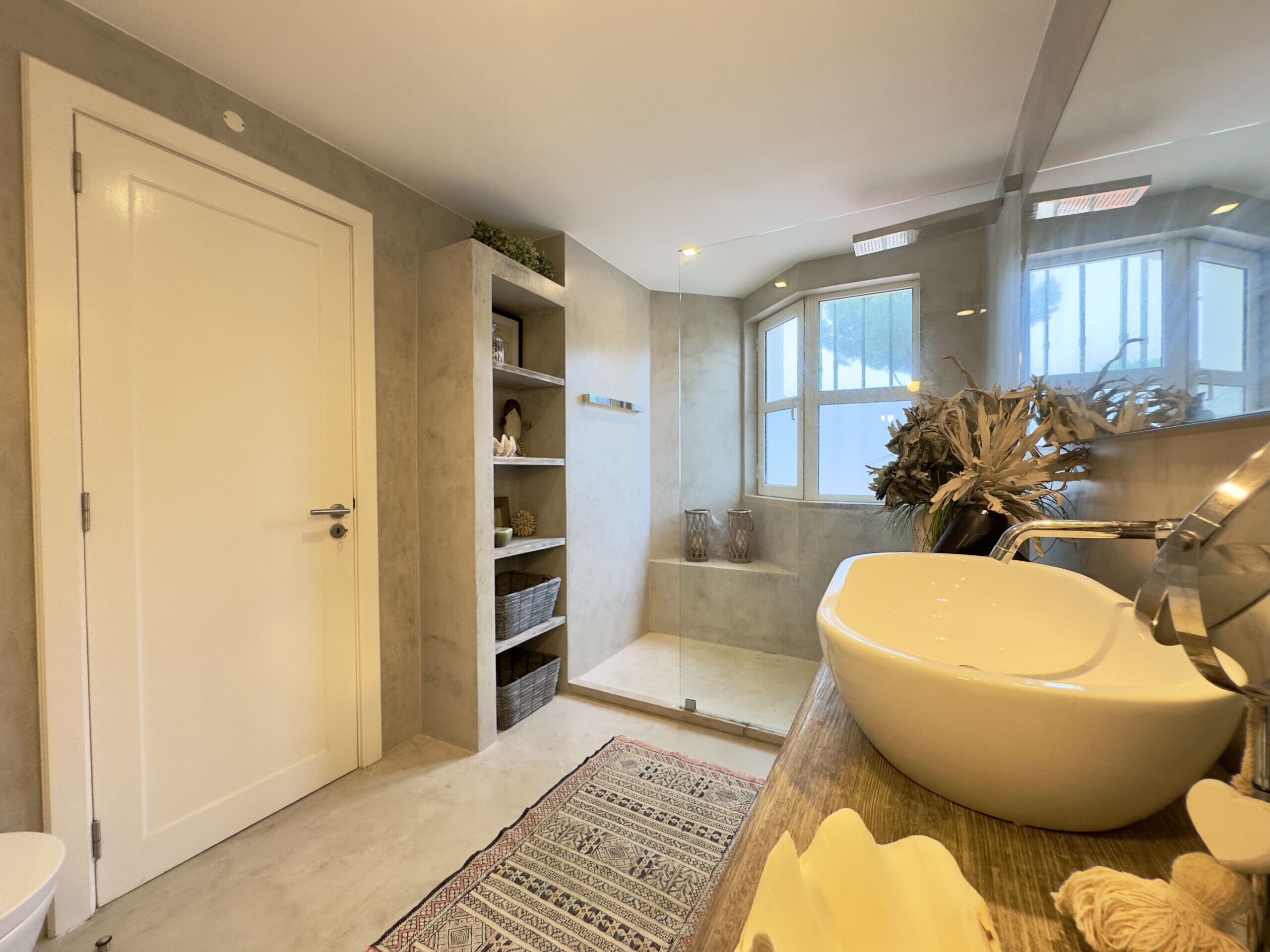 A modern bathroom with a large sink, shelves with decorative items, a window, and a door.