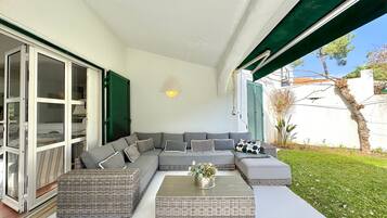Een overdekt terras met een bank en twee bijzettafeltjes, een schuifpui en een groen zonnescherm.