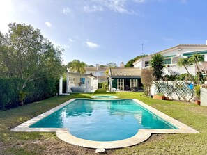 Ein Pool mit geschwungener Kante, umgeben von einer gepflegten Rasenfläche und verschiedenen Pflanzen. Im Hintergrund sind zwei Häuser mit Fenstern und einer Satellitenschüssel zu sehen.