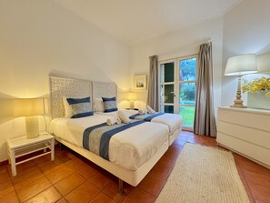 Ein Schlafzimmer mit einem großen Bett, zwei Nachttischen, einem Schreibtischchen, einem Fenster mit Vorhängen und einer Lampe auf einer Kommode.