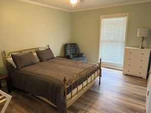 Une chambre à coucher avec un lit à ossature métallique, une chaise, une commode et une fenêtre avec des stores.