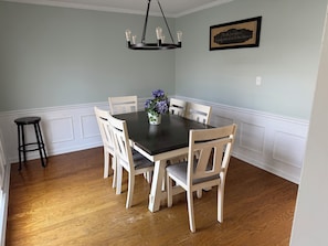 Une salle à manger avec une table en bois foncé, des chaises blanches, un lustre et une citation encadrée accrochée au mur.