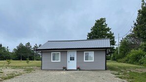 A small, single-story building with a grey exterior and a white door, surrounded by a gravel driveway and grassy areas. - Oakhill Oasis Guest House (Oak Hill)