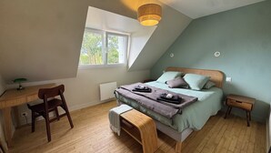 Ein Schlafzimmer mit schrägverdachter Decke, einem Bett mit Kissen, einem Schreibtisch mit Stuhl, einem kleinen Nachttisch und einem Fenster mit Blick auf Bäume.