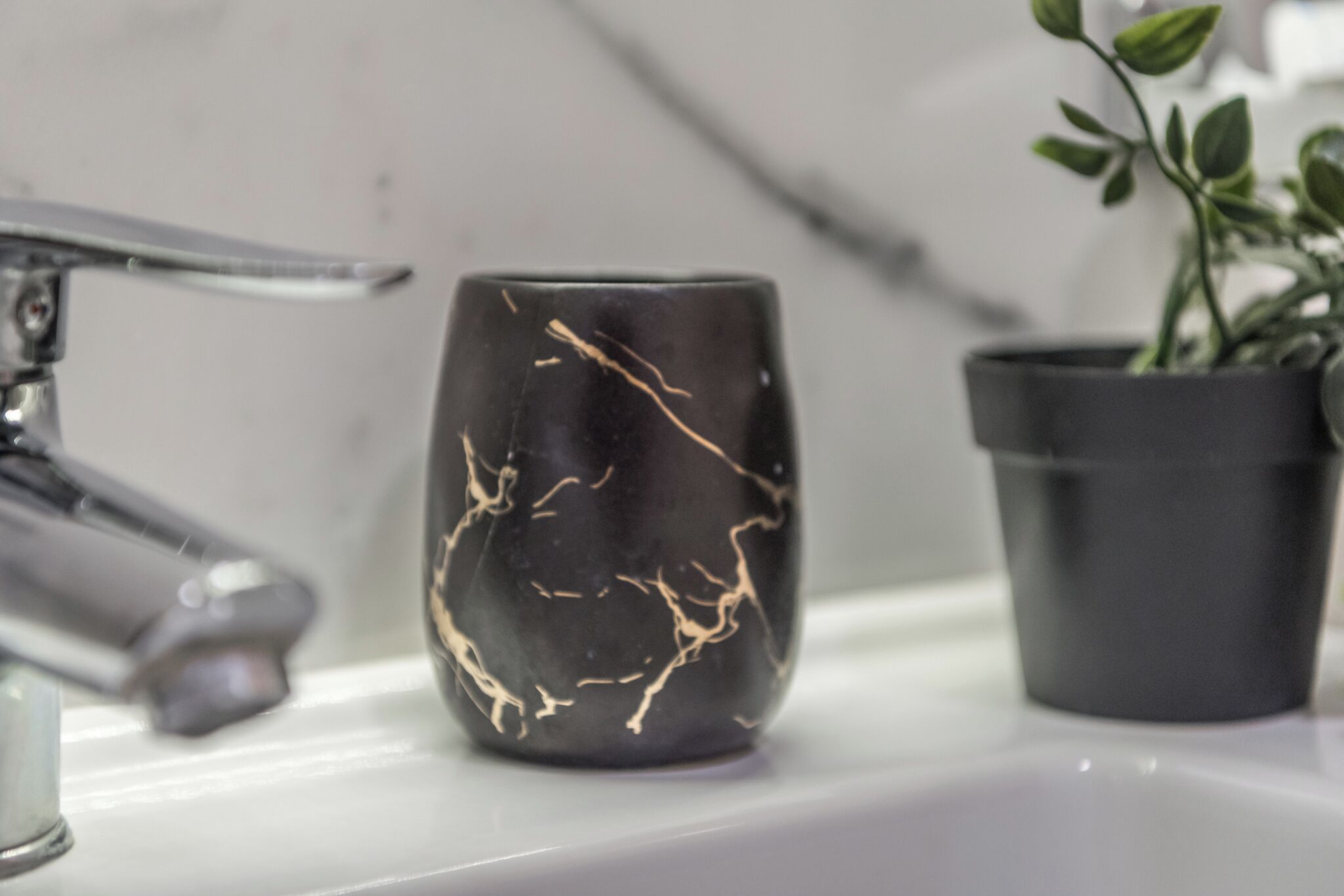 Une tasse en marbre noir aux veines dorées, posée sur un comptoir blanc, avec un robinet moderne et une plante en pot.