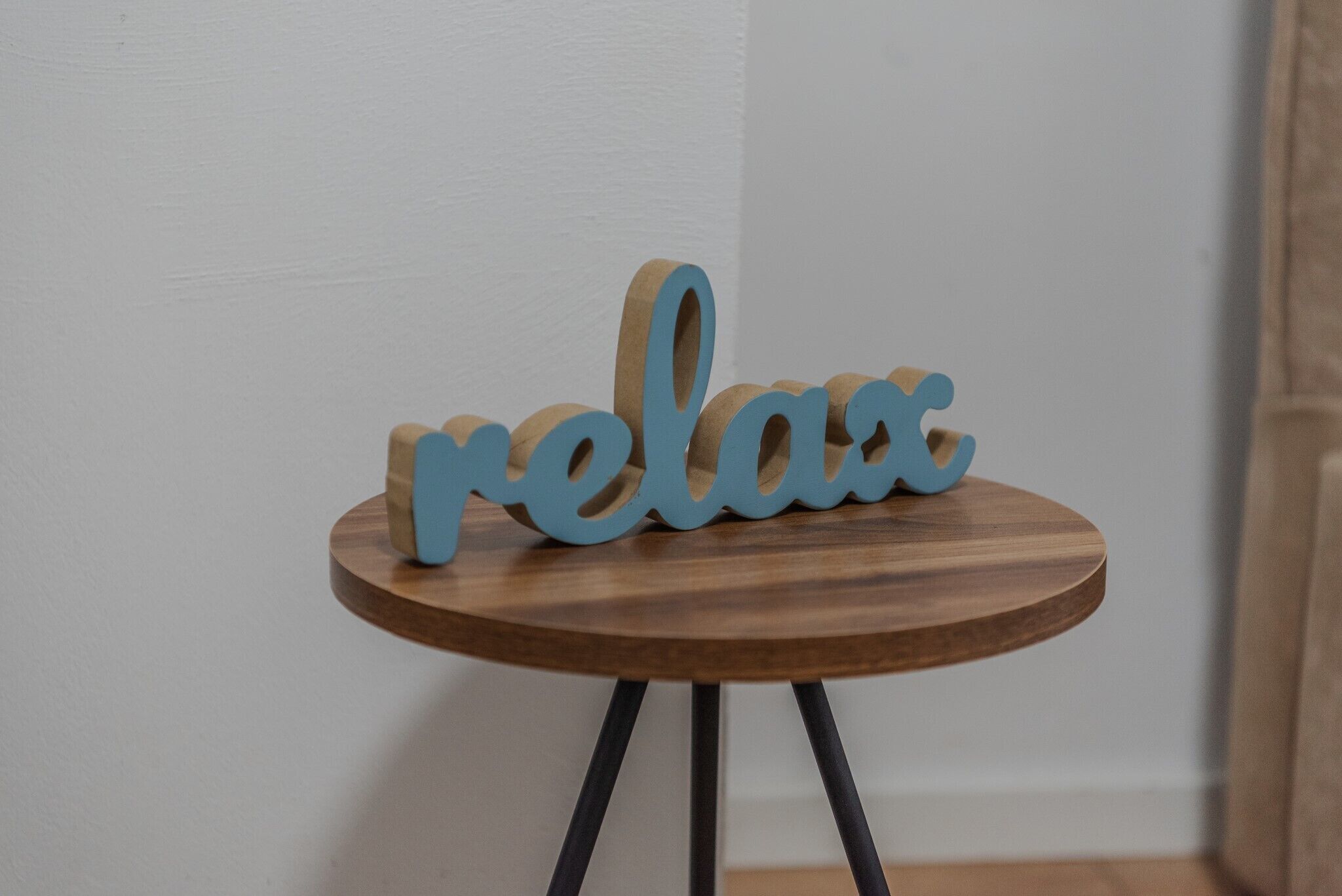 Une table en bois, dotée d’un plateau rond en bois et de pieds noirs, sur laquelle est posé un panneau en bois portant le mot « relax » en lettres bleues.