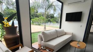 A modern living room with a sofa, two armchairs, a coffee table, and a small side table. There is a large window with a view of a garden and palm trees. A flat-screen TV is mounted on the wall. - pousada ibiza (Pirenopolis)