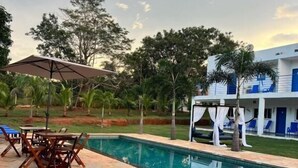 A swimming pool with a tiled deck, a covered seating area, and a building with blue accents in the background. - pousada ibiza (Pirenopolis)