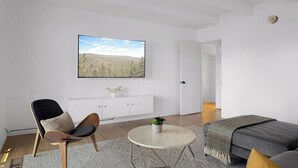 Ein modernes Wohnzimmer mit einem großen Flachbildfernseher, einer Couch, einem Sessel, einem kleinen runden Tisch und einem Schrank.