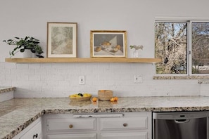 A kitchen with white brick walls, a granite countertop, two framed pictures, a potted plant, and a window with a view of trees. - Modern Lakeview Retreat, Beach Club Access with 2 Decks (Lake Arrowhead)