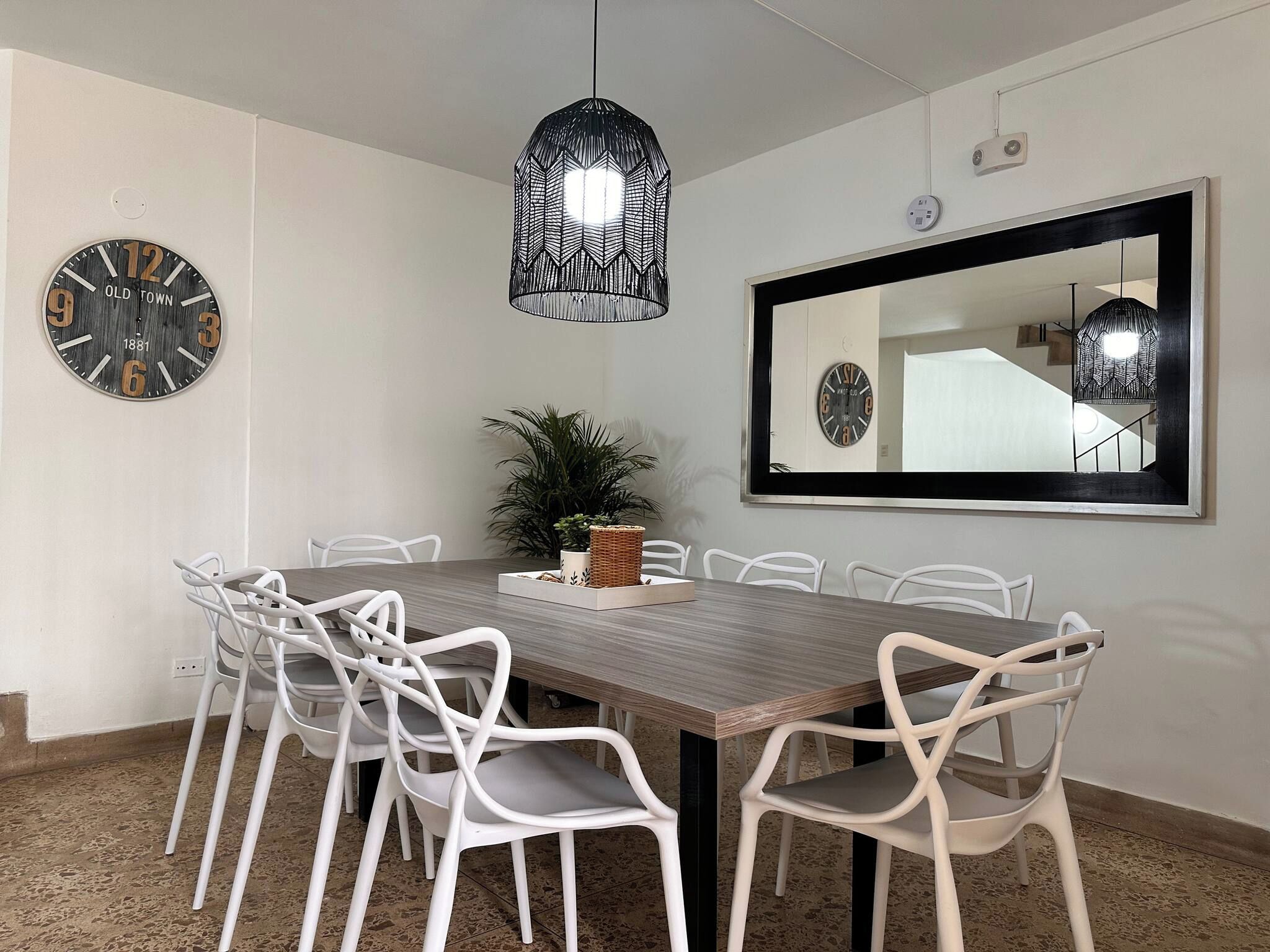 A dining area with a wooden table, white chairs, a hanging pendant light, a potted plant, and two wall clocks.